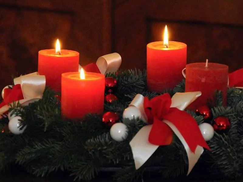Vijf rode kaarsen branden op een kerstkrans met groene takken en versiering.