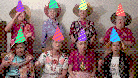 50 jaar man humor gif - Een groep oudere vrouwen in feesthoeden, zingend en blij in een woonkamer.