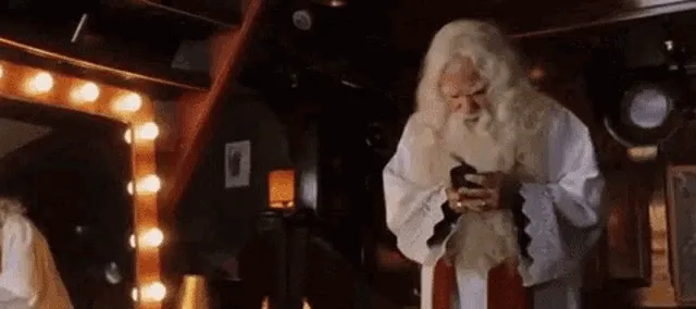 Een oudere man met een lange witte baard kijkt naar zijn telefoon in een gezellige ruimte.